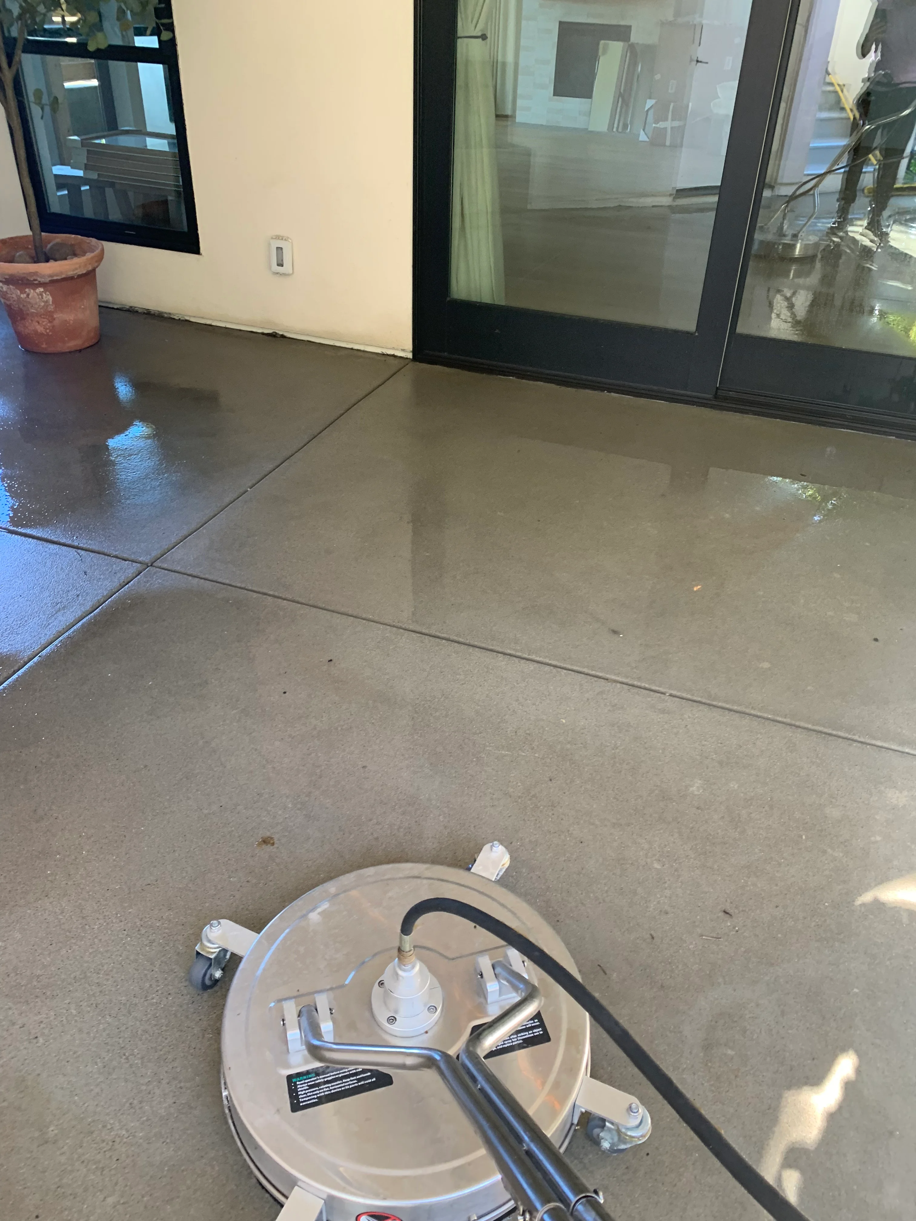 Concrete patio cleaning in progress with pressure washing equipment and glass doors visible